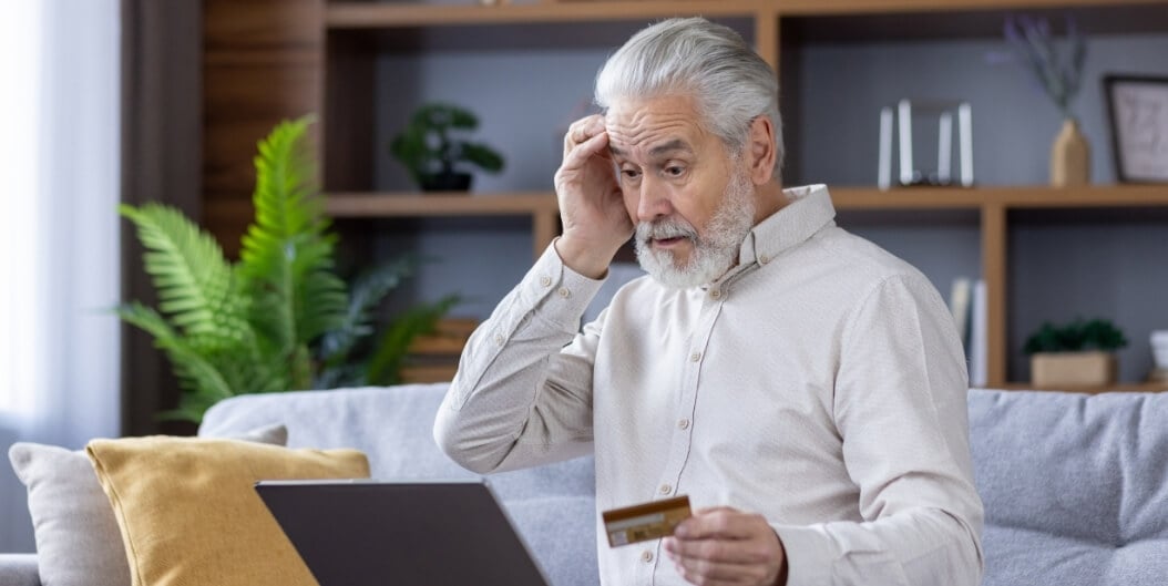 Elderly Man Holding a Credit Card and Looking Stressed While Using a Laptop at an Independent Living Facility