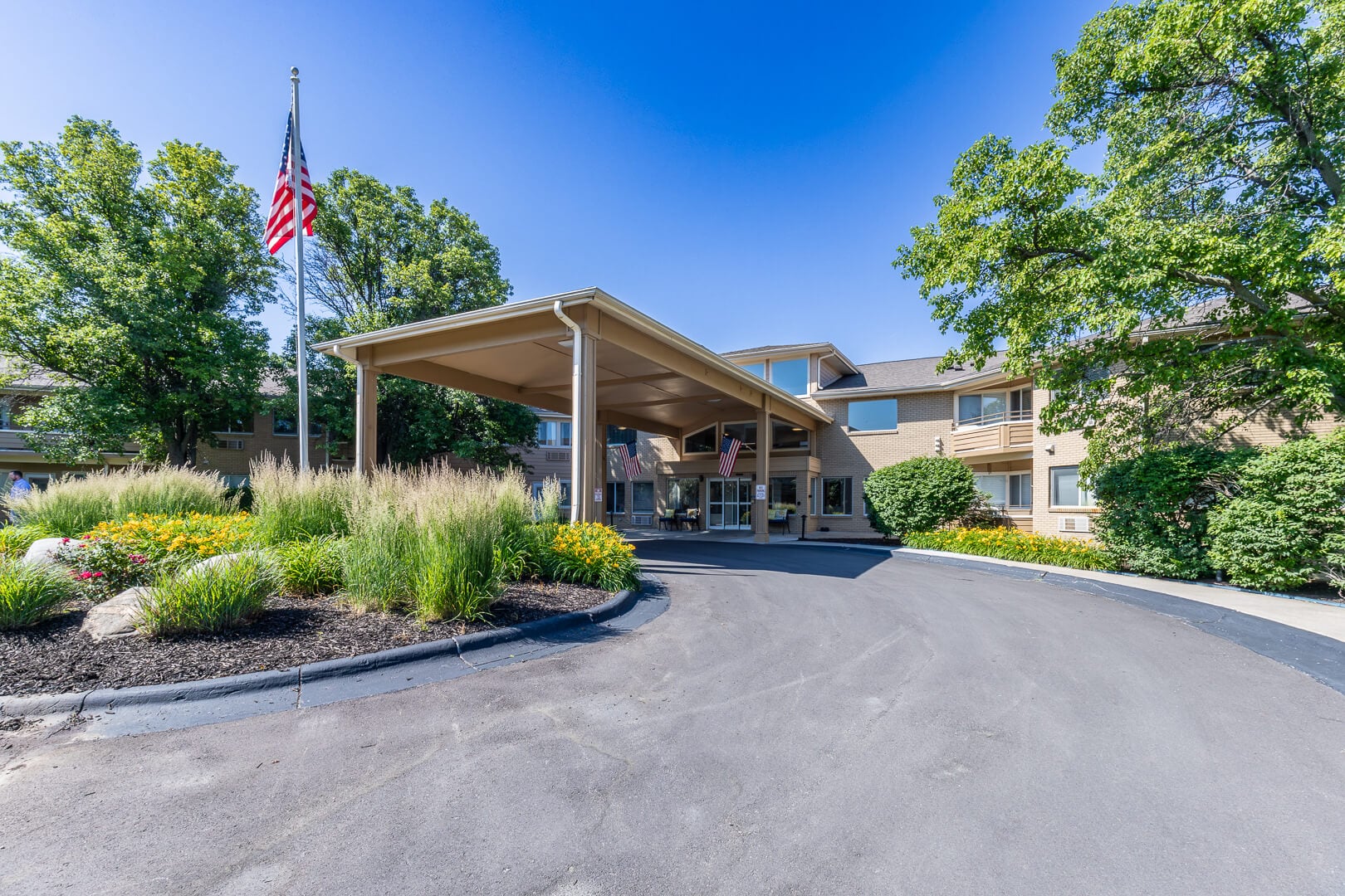 Tree-Lined Front Entrance with Covered Porte-Cochere and American Flag at Independent Living Community